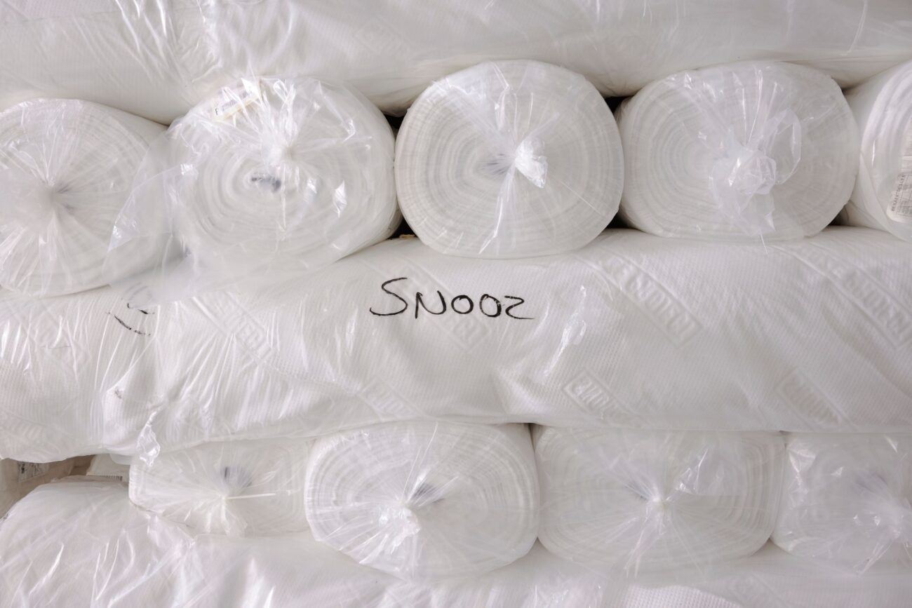 Mehrere große weiße, in Plastik eingewickelte Stoffrollen sind übereinander gestapelt. Auf einer Rolle in der Mitte steht handschriftlich das Wort SNOOZ auf der Plastikhülle.