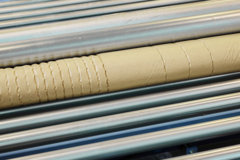 Nahaufnahme einer Papphülse mit gleichmäßig verteilten Rillen, die auf parallelen Metallrollen in einer Produktionsstätte ruht, die sich der Verantwortung und dem Schutz der Umwelt verschrieben hat.