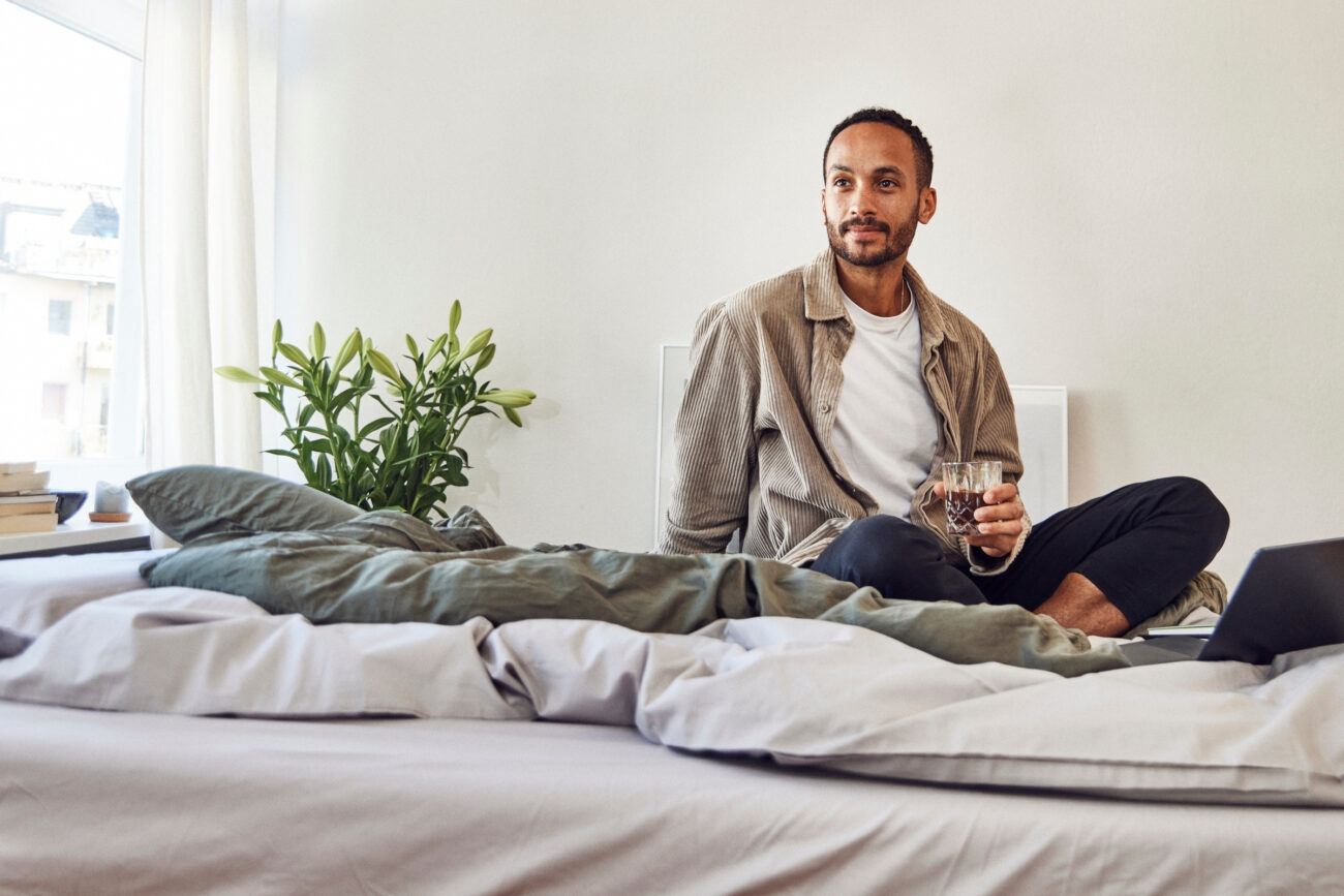 Ein Mann sitzt im Schneidersitz auf einem Bett, hält ein Glas in der Hand und blickt nachdenklich zur Seite. In einem hellen, gemütlichen Raum stehen eine Pflanze, Bücher und ein Laptop auf oder neben dem Bett.