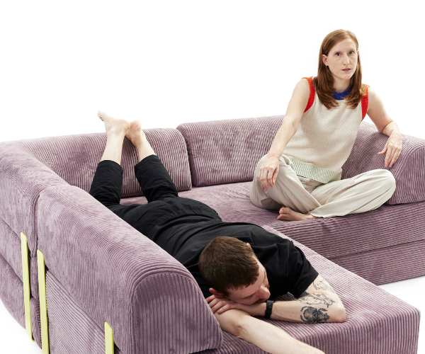 Eine Frau sitzt aufrecht auf einem hellvioletten Sektionssofa und blickt nach vorne, während ein Mann mit dem Gesicht nach unten und ausgestreckten Beinen auf demselben Sofa liegt. Der Hintergrund ist einfarbig weiß.