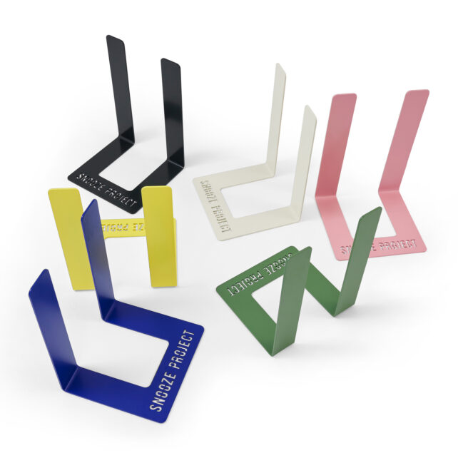 Fünf metallene Buchstützen in Schwarz, Gelb, Weiß, Rosa, Blau und Grün sind auf einer weißen Fläche angeordnet. Auf jeder Buchstütze ist das SNOOZE PROJECT ausgeschnitten.