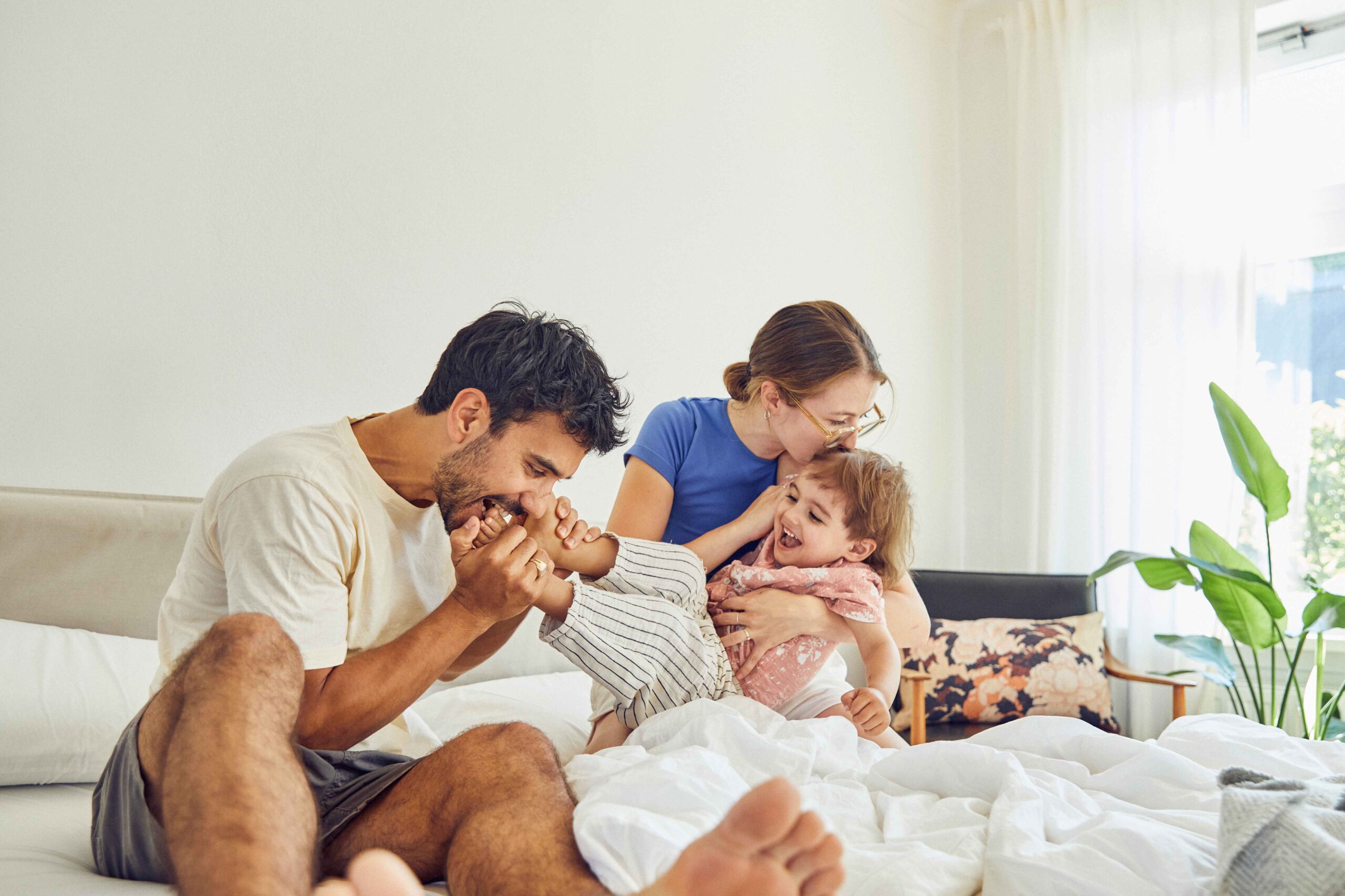 Eine glückliche vierköpfige Familie sitzt auf einem Familienbett, lacht und spielt zusammen. Der Vater hält spielerisch den Fuß eines Kindes, während die Mutter ein anderes lächelndes Kind umarmt und küsst. Sonnenlicht strömt durch das Fenster in diese gemütliche Erlebniswelt.