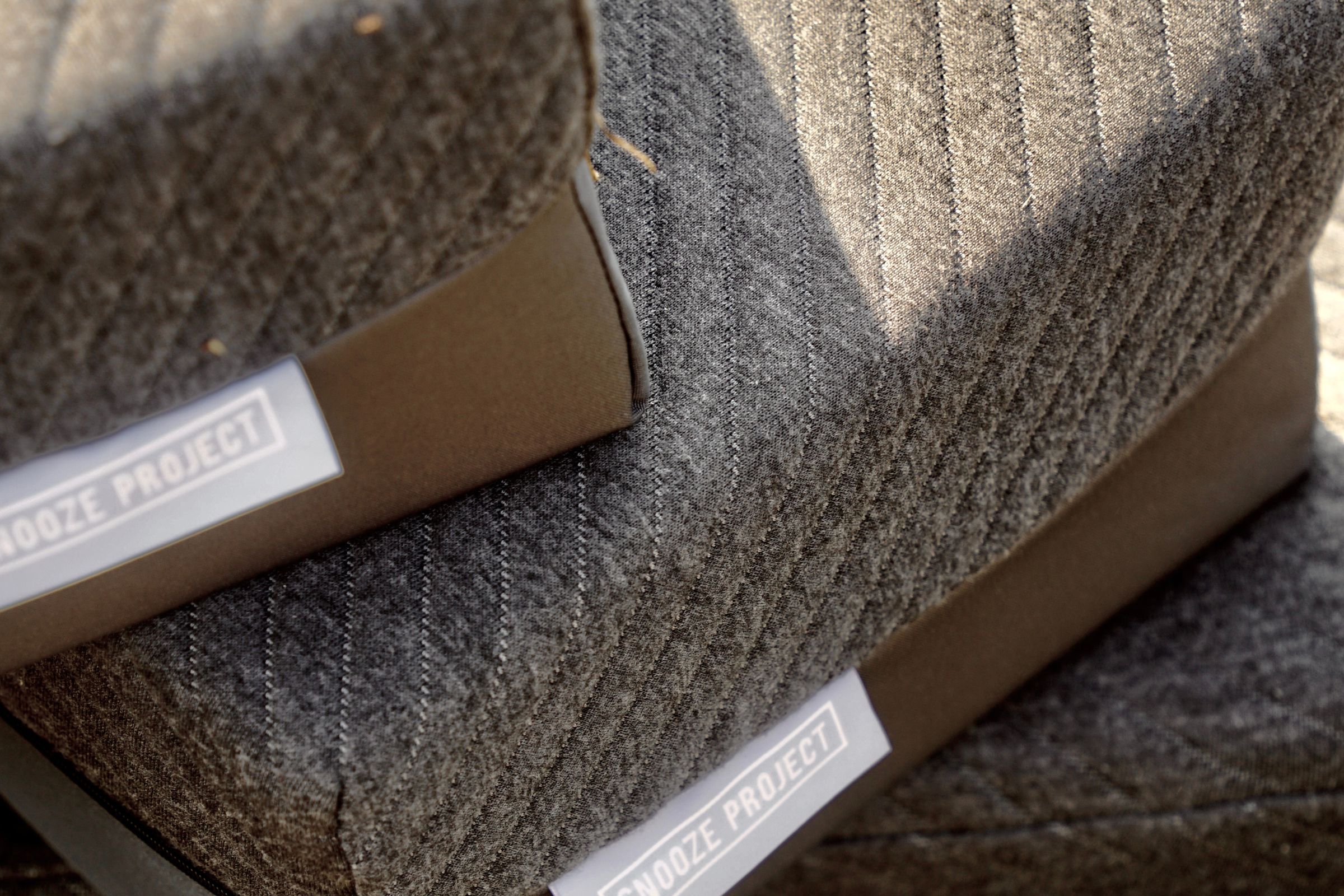 Nahaufnahme von zwei grauen, strukturierten, gepolsterten Kissen mit diagonalen Nähten und der Aufschrift "Snooze Project" auf weißen Schildern - perfekt für Ihre Erlebniswelt oder in Kombination mit Matratzen nach Maß.