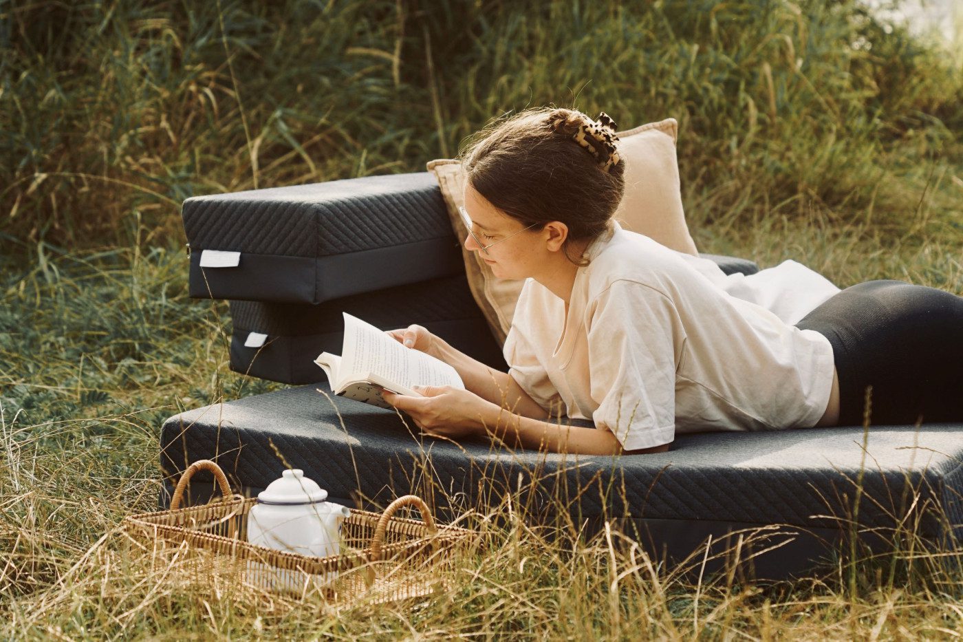 Eine Frau liegt auf einem Stapel Matratzen nach Maß im Freien und liest ein Buch. Auf einer Wiese, mit Picknickkorb und Teekanne in der Nähe, genießt sie einen friedlichen Moment in der Natur - ihre eigene kleine Erlebniswelt.