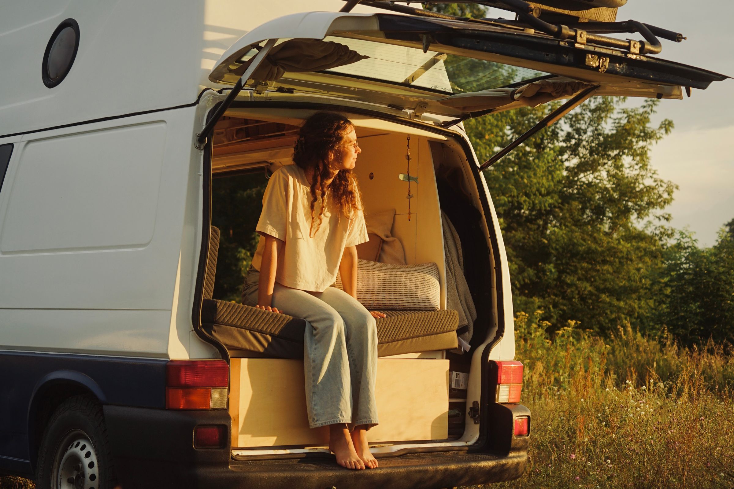 Eine Frau sitzt barfuß auf dem offenen Rücksitz eines weißen Wohnmobils und blickt dem Sonnenlicht entgegen. Umgeben von Natur und Wildblumen genießt sie ein gemütliches Interieur mit Matratzen nach Maß für ultimativen Komfort - ihre eigene private Erlebniswelt auf Rädern.