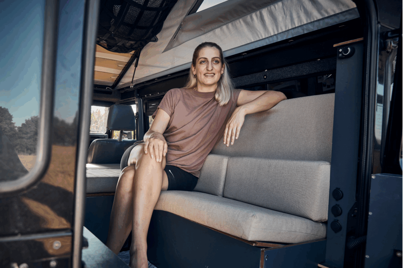 Eine Person mit langen blonden Haaren sitzt lächelnd auf einem grauen Polster nach Maß im Inneren eines Wohnmobils, während das Sonnenlicht durch die offene Tür fällt und draußen Bäume zu sehen sind.