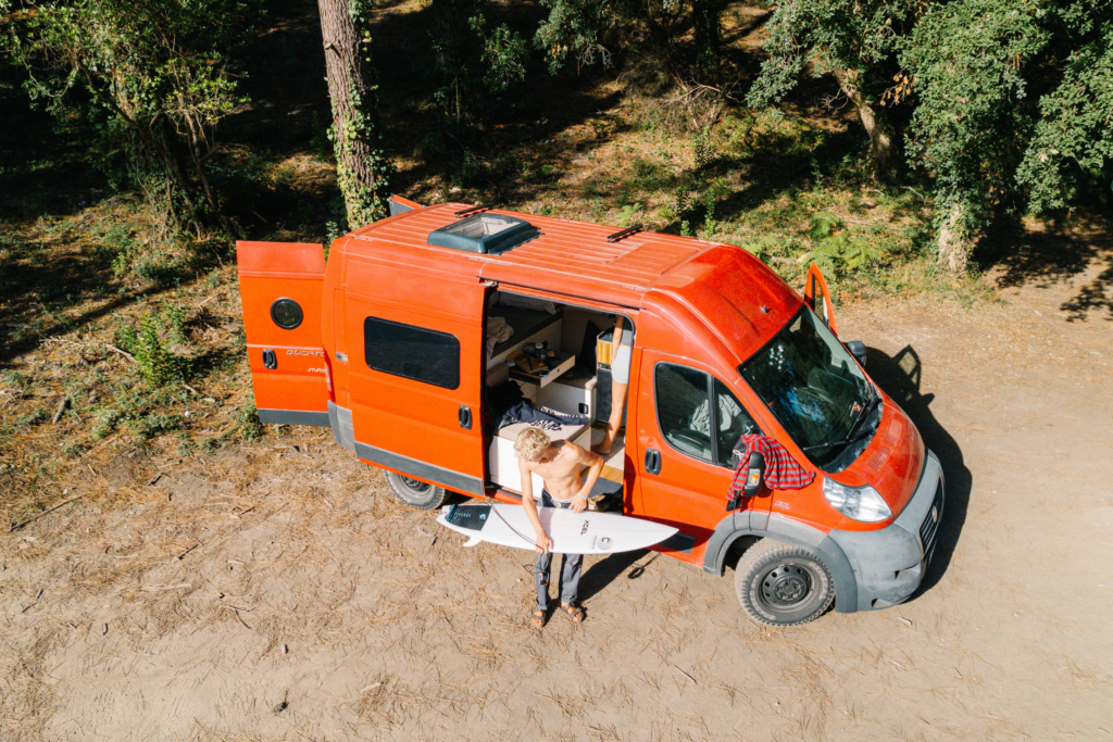 Eine Person steht mit einem Surfbrett in der Hand vor einem roten Wohnmobil mit geöffneter Seitentür in einem sonnigen Waldgebiet. Im Inneren schaffen individuelles Campingzubehör und Polster nach Maß eine gemütliche Erlebniswelt für Outdoor-Abenteuer.