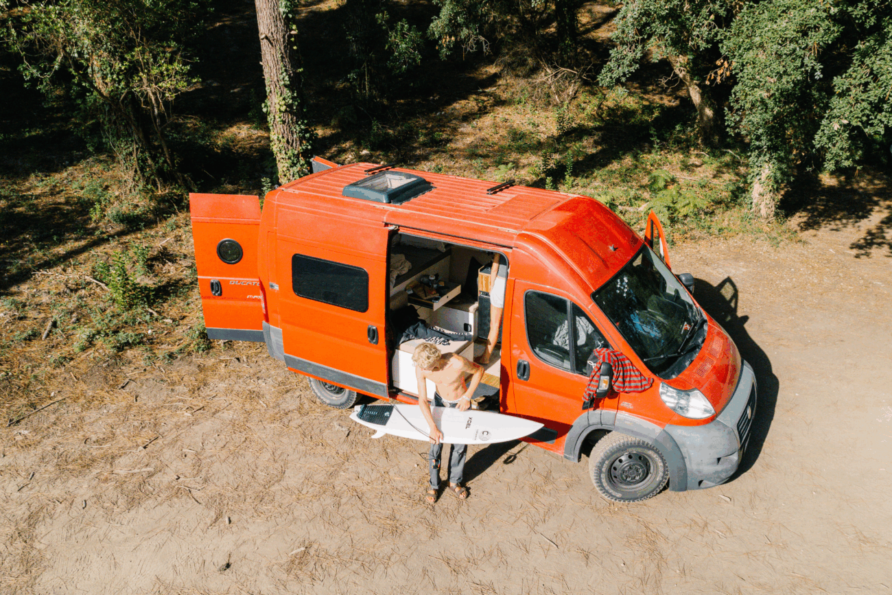Eine Person steht mit einem Surfbrett in der Hand vor einem roten Wohnmobil mit geöffneter Seitentür in einem sonnigen Waldgebiet. Im Inneren schaffen individuelles Campingzubehör und Polster nach Maß eine gemütliche Erlebniswelt für Outdoor-Abenteuer.