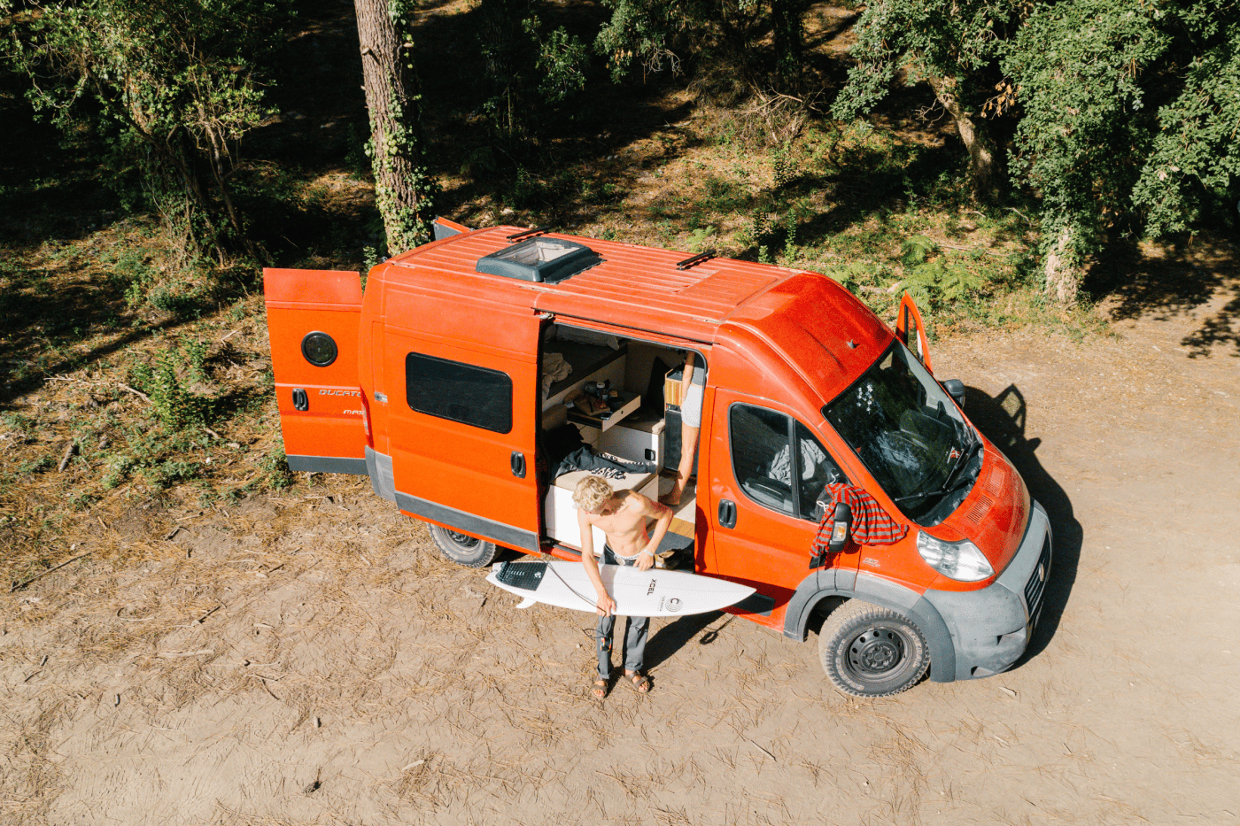 Eine Person steht mit einem Surfbrett in der Hand vor einem roten Wohnmobil mit geöffneter Seitentür in einem sonnigen Waldgebiet. Im Inneren schaffen individuelles Campingzubehör und Polster nach Maß eine gemütliche Erlebniswelt für Outdoor-Abenteuer.