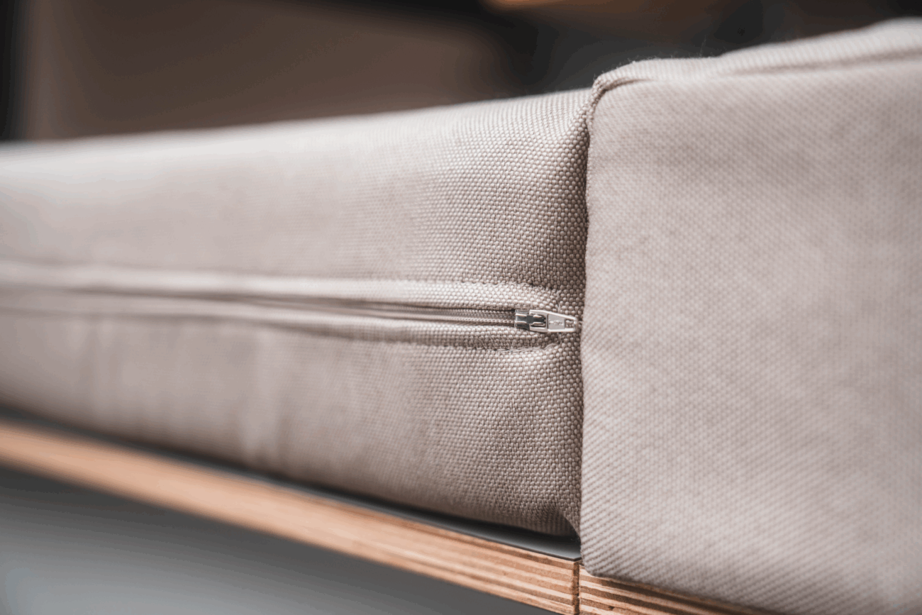 Nahaufnahme eines beigen Stoffkissens mit sichtbarem Reißverschluss an der Seite, das auf einer Holzoberfläche liegt - die perfekte Inspiration für Ihr eigenes Polster nach Maß mit unserem Konfigurator. Die Textur und die Details des Reißverschlusses sind deutlich zu erkennen.