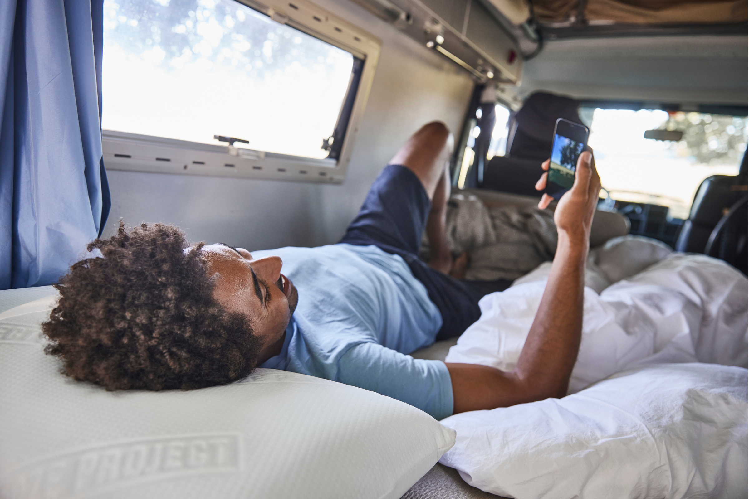 Eine Person liegt auf einem Bett mit Polster nach Maß in einem Wohnmobil, hält ein Smartphone hoch und schaut es an. Das Sonnenlicht fällt durch das Fenster, und um sie herum sind Kissen und Bettwäsche zu sehen.