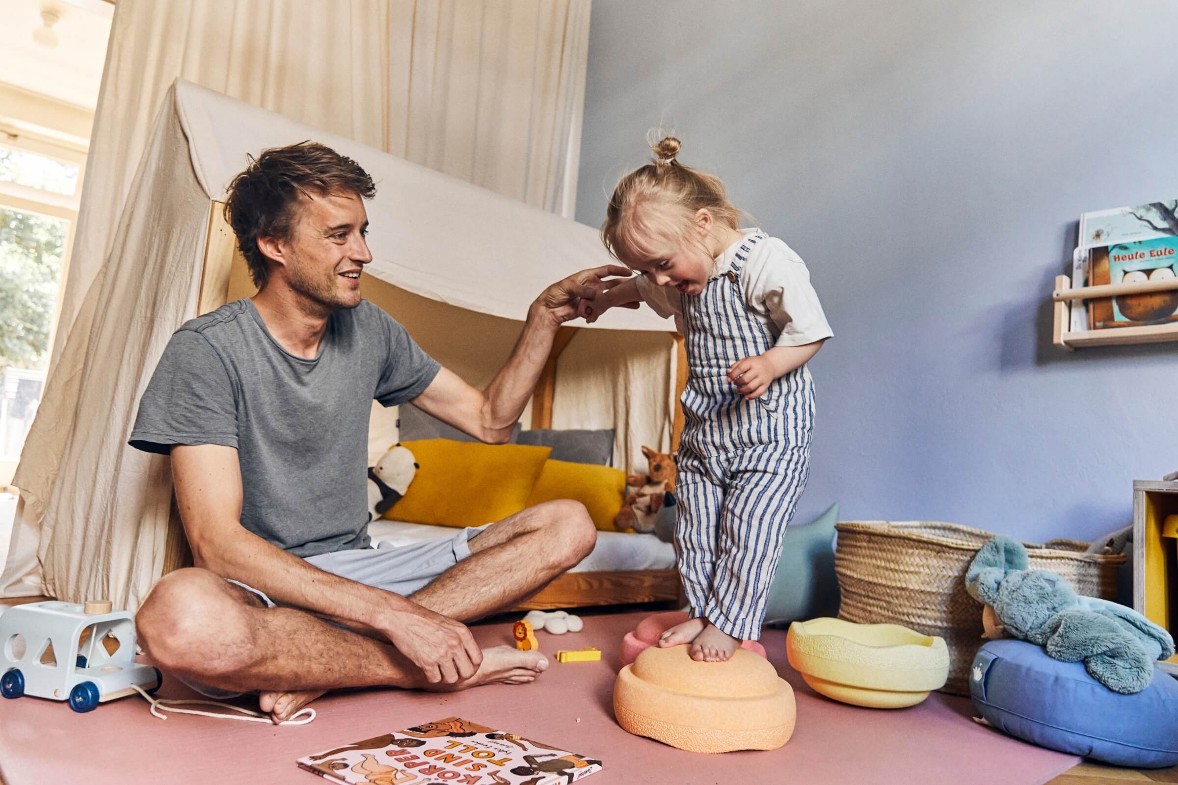 Ein Mann sitzt im Schneidersitz auf dem Boden, lächelt und streckt die Hand nach einem kleinen Kind aus, das auf einer weichen Matratze steht. Sie spielen zusammen in einer gemütlichen, mit Spielzeug gefüllten Erlebniswelt mit Büchern und Plüschtieren im Hintergrund.