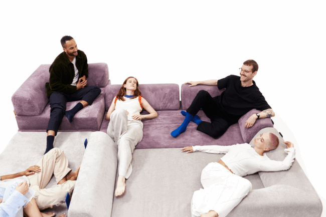 Vier Personen entspannen sich auf einem großen, hellvioletten Schlafsofa in einem weißen Raum, wobei zwei aufrecht sitzen und zwei liegen und sich zu unterhalten und die Gesellschaft des anderen zu genießen scheinen. Perfekt für jede Kategorieseite, die auf Komfort und Stil setzt.