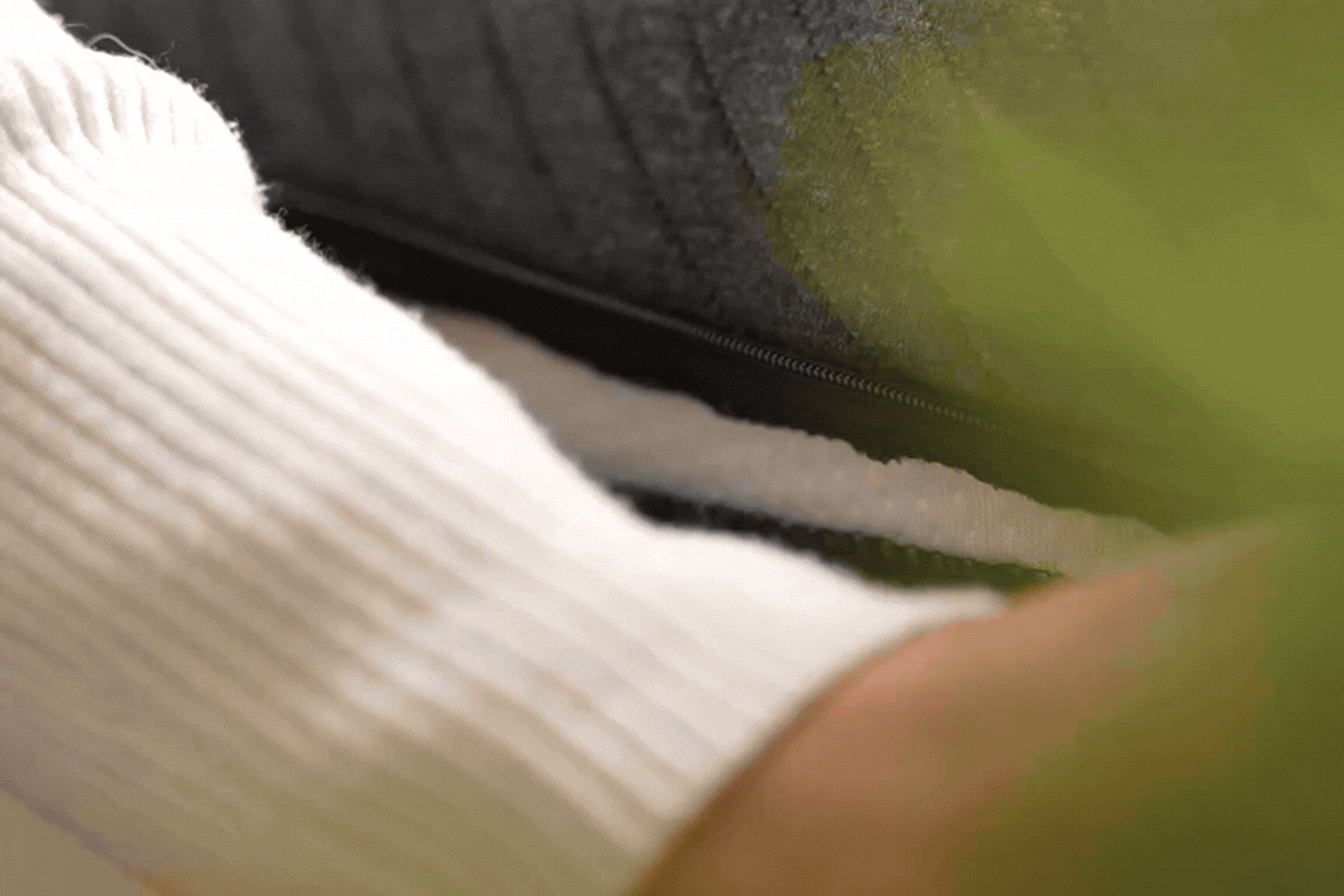 Eine Person, die einen weißen gerippten Pullover trägt, zieht einen schwarzen Reißverschluss an einem dunklen Stoffkissen oder Polstermöbel auf, um die Erlebniswelt individueller Matratzen nach Maß zu erkunden, wobei im Vordergrund etwas Grün zu sehen ist.
