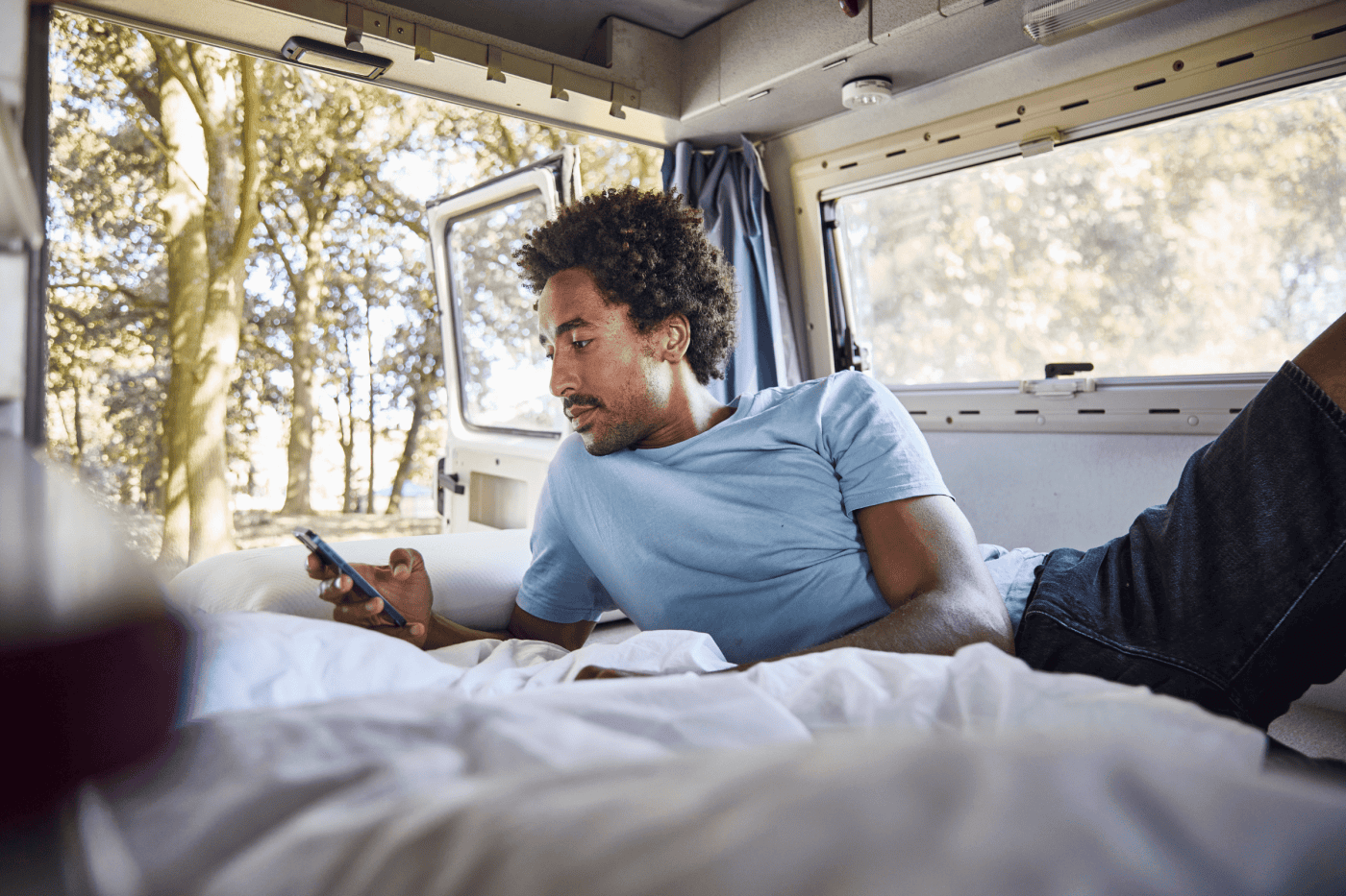 Ein Mann liegt auf einem Bett in einem Wohnmobil und schaut auf sein Smartphone. Das Sonnenlicht fällt durch große Fenster ein, und draußen sind Bäume zu sehen. Er wirkt entspannt, trägt ein hellblaues Hemd und Jeans.