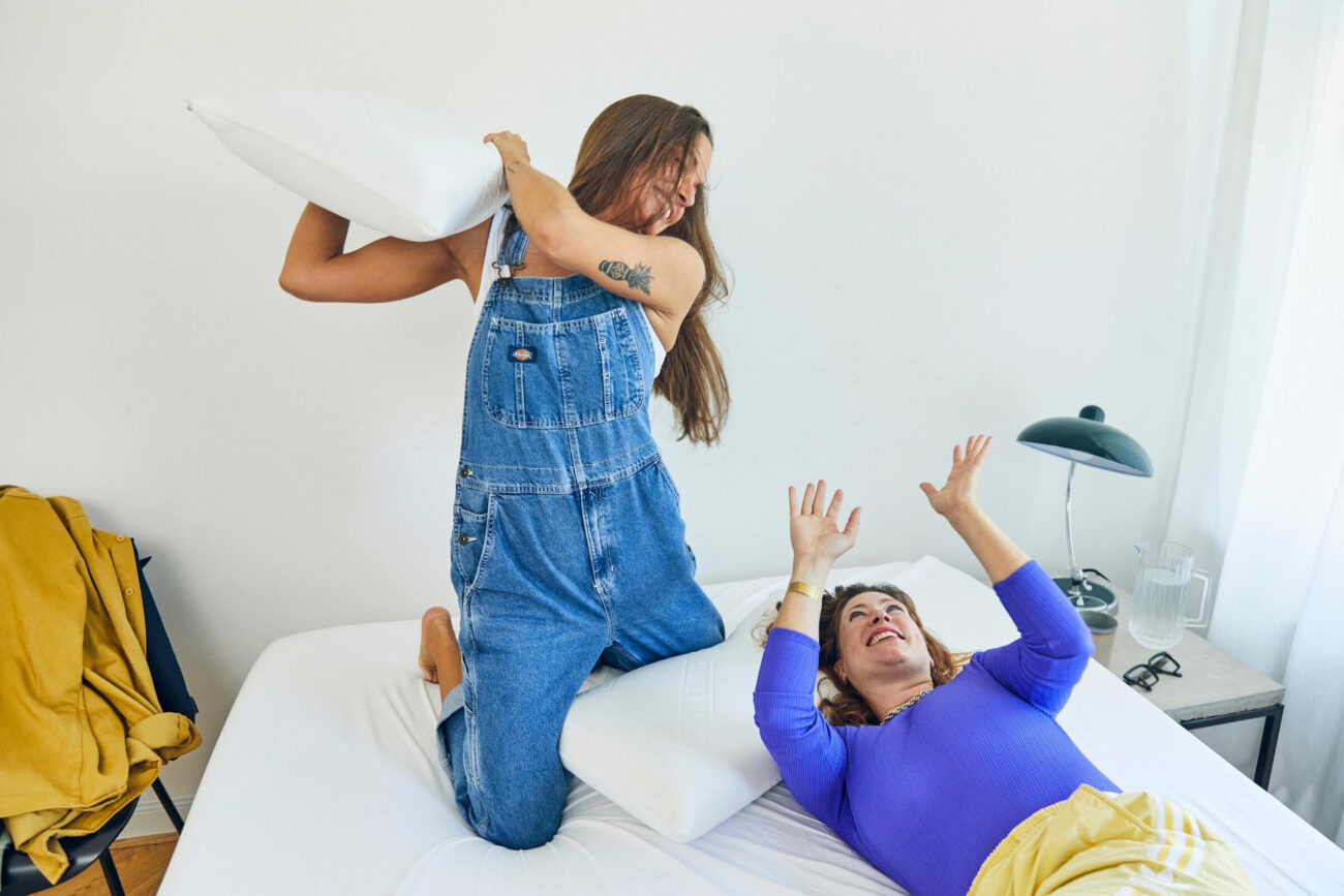 Zwei Frauen bei einer spielerischen Kissenschlacht auf einem Bett, beide lächelnd in einem hellen, gemütlichen Schlafzimmer - entdecken Sie pure Freude in Ihrer Erlebniswelt mit Matratzenschoner nach Maß, der alles bequem und geschützt hält.