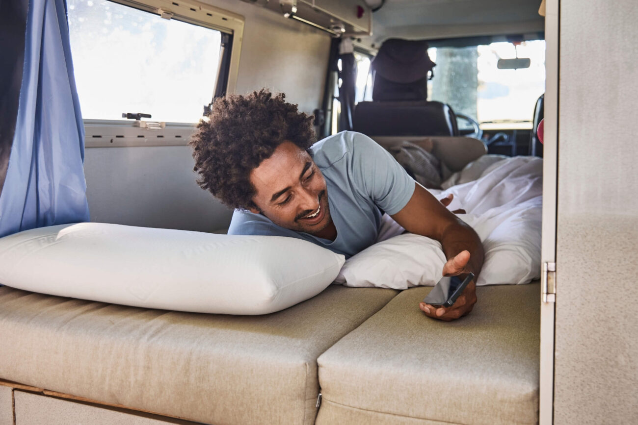 Ein Mann liegt auf einem Bett in einem Wohnmobil und schaut lächelnd auf sein Telefon, umgeben von Kissen und Bettzeug. Natürliches Licht fällt durch das Fenster neben ihm, wo ein Konfigurator Matratzenschoner nach Maß seine Matratze schützt.