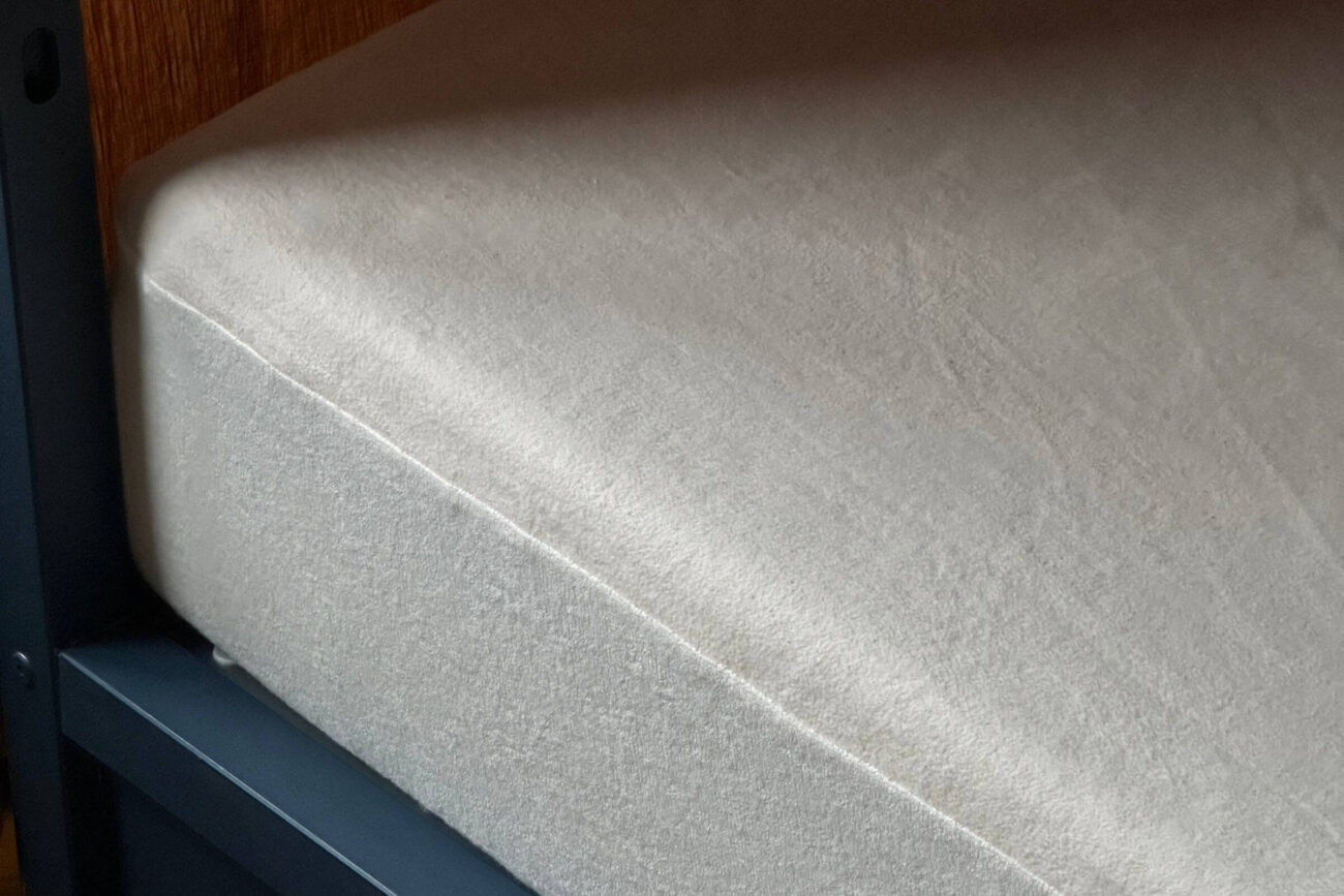 Eine Nahaufnahme eines ordentlich gemachten Bettes mit einem einfarbigen, hellen Spannbettlaken und einem Matratzenschoner nach Maß, der die Matratze bedeckt. Das Bettgestell ist dunkel, und ein Teil einer Holzwand trägt zur Erlebniswelt im Hintergrund bei.