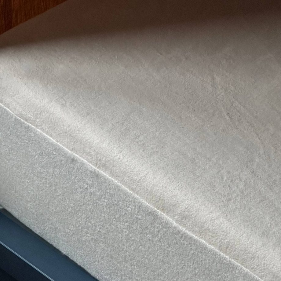 Eine Nahaufnahme eines ordentlich gemachten Bettes mit einem einfarbigen, hellen Spannbettlaken und einem Matratzenschoner nach Maß, der die Matratze bedeckt. Das Bettgestell ist dunkel, und ein Teil einer Holzwand trägt zur Erlebniswelt im Hintergrund bei.