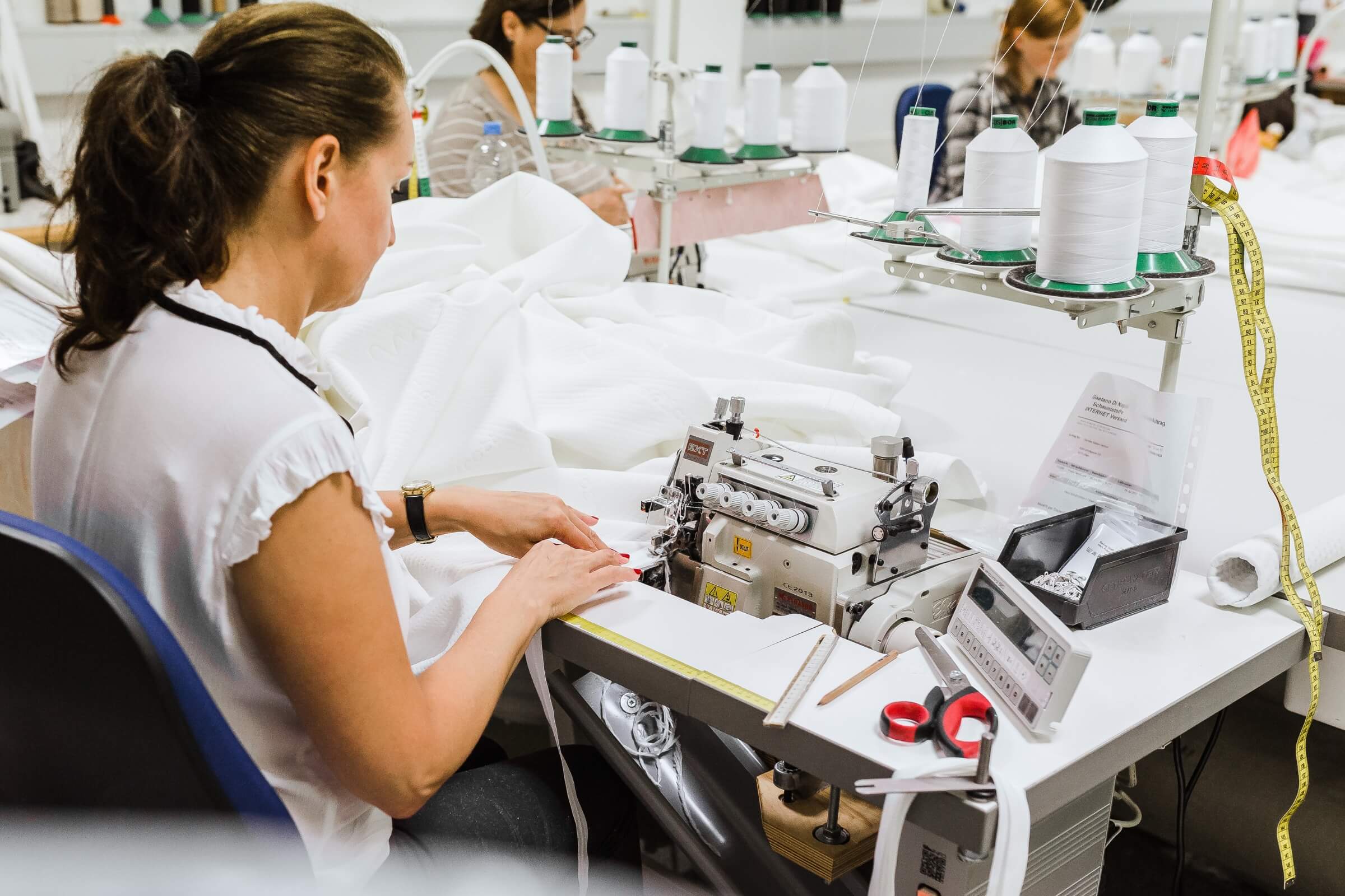 Eine Frau bedient eine Nähmaschine an einem Arbeitsplatz in einer Textilfabrik, umgeben von weißem Stoff und Werkzeugen, und stellt Spannbettlaken nach Maß her. Im Hintergrund sind andere Arbeiter zu sehen, die ebenfalls nähen.