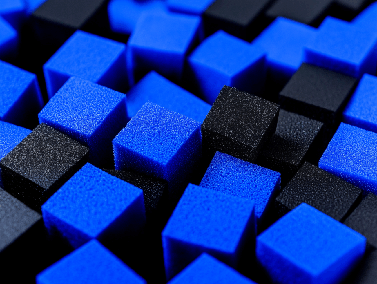 Eine Nahaufnahme von blauen und schwarzen Filterschaumwürfeln, die in einem Gittermuster angeordnet sind, wodurch eine strukturierte und geometrische Oberfläche entsteht.