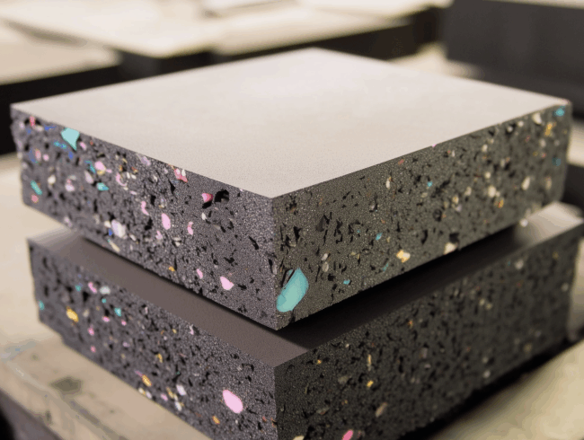 Nahaufnahme von zwei gestapelten, dicken schwarzen Filterschaum-Matten mit eingebetteten bunten Flecken, die auf einer flachen Oberfläche in einem Innenraum liegen.