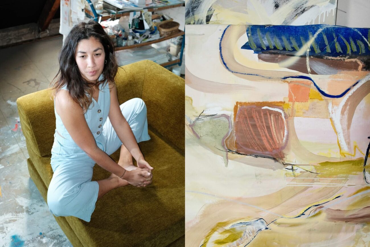 Eine Frau in einem hellblauen Overall sitzt barfuß auf einer grünen Couch in einem Künstleratelier; neben ihr ist ein abstraktes Gemälde mit geschwungenen Linien und Farbflecken in Blau, Braun und Pfirsich zu sehen.