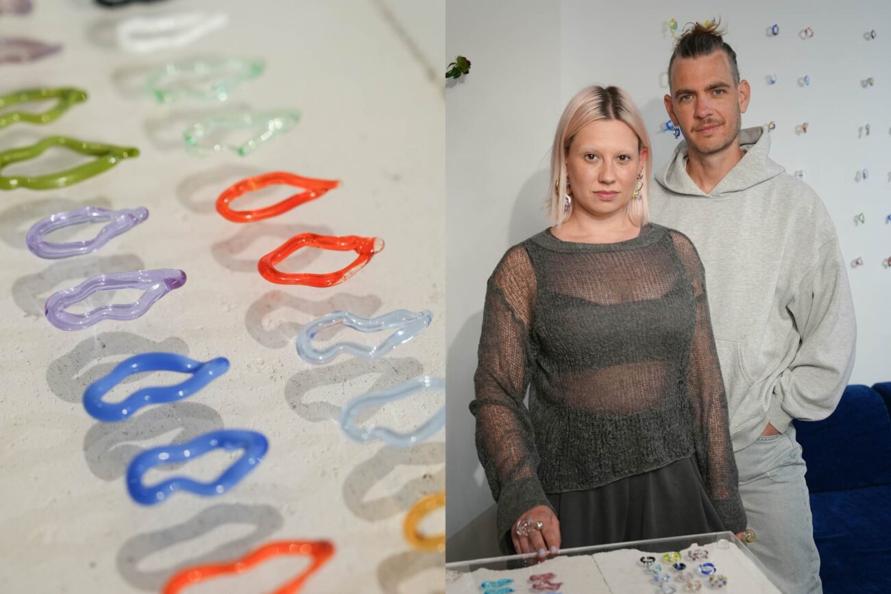 Auf der linken Seite sind bunte Glasohrringe in Form von Lippen auf einer hellen Fläche ausgestellt. Rechts stehen eine Frau und ein Mann gemeinsam vor einer Wand, auf einem Tisch vor ihnen sind einige Schmuckstücke zu sehen.