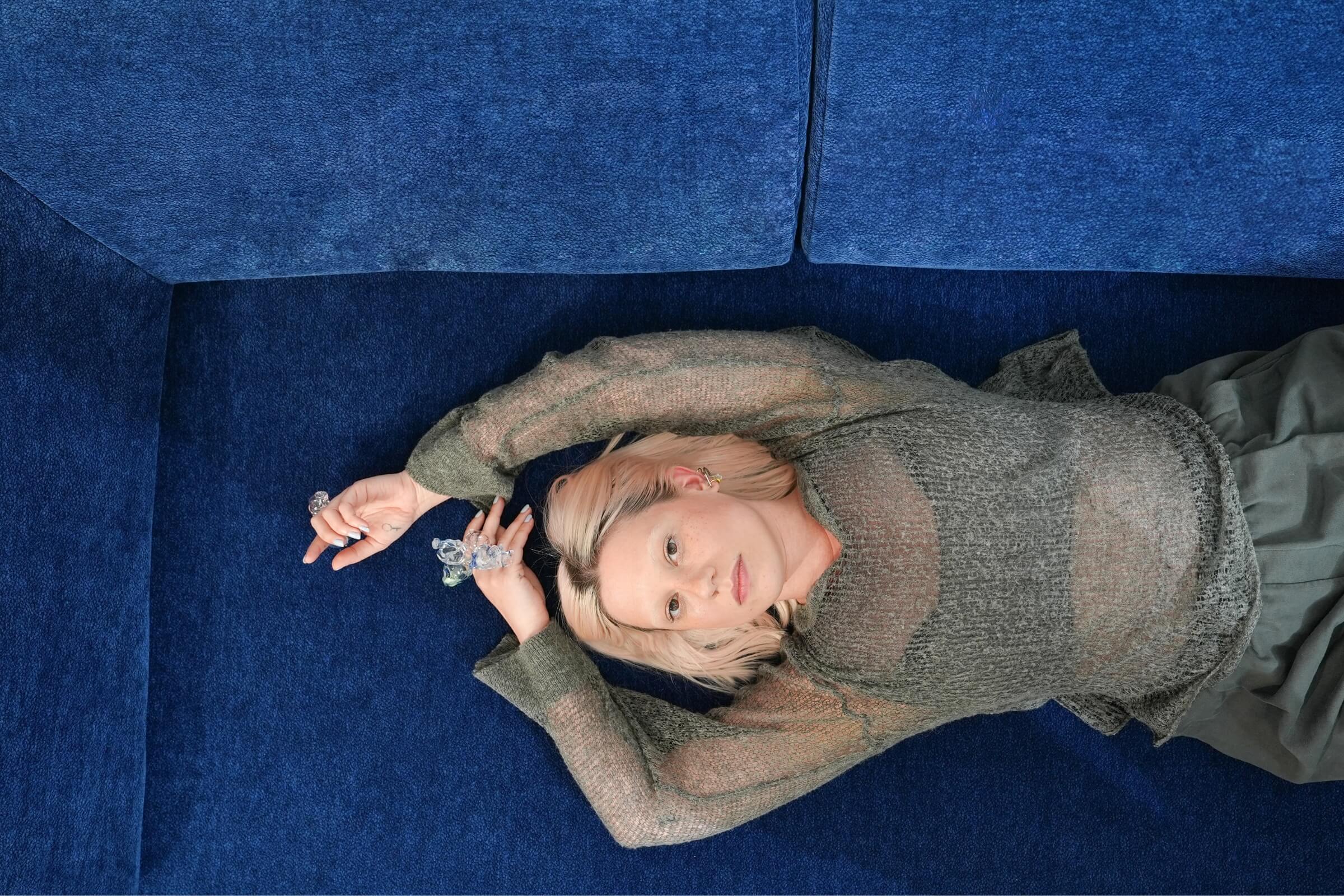 Eine Person mit blondem Haar liegt auf einer dunkelblauen Couch und blickt mit über den Kopf gestreckten Armen in die Kamera. Sie trägt ein durchsichtiges, langärmeliges Oberteil und einen grauen Rock.
