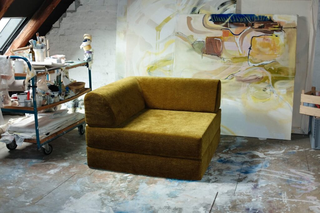 Ein senfgelber, moderner Sessel steht im Atelier eines Künstlers, umgeben von Kunstzubehör, einem Rollwagen und einem abstrakten Gemälde, das an einer weißen Backsteinwand mit einem mit Farbe bespritzten Boden lehnt.