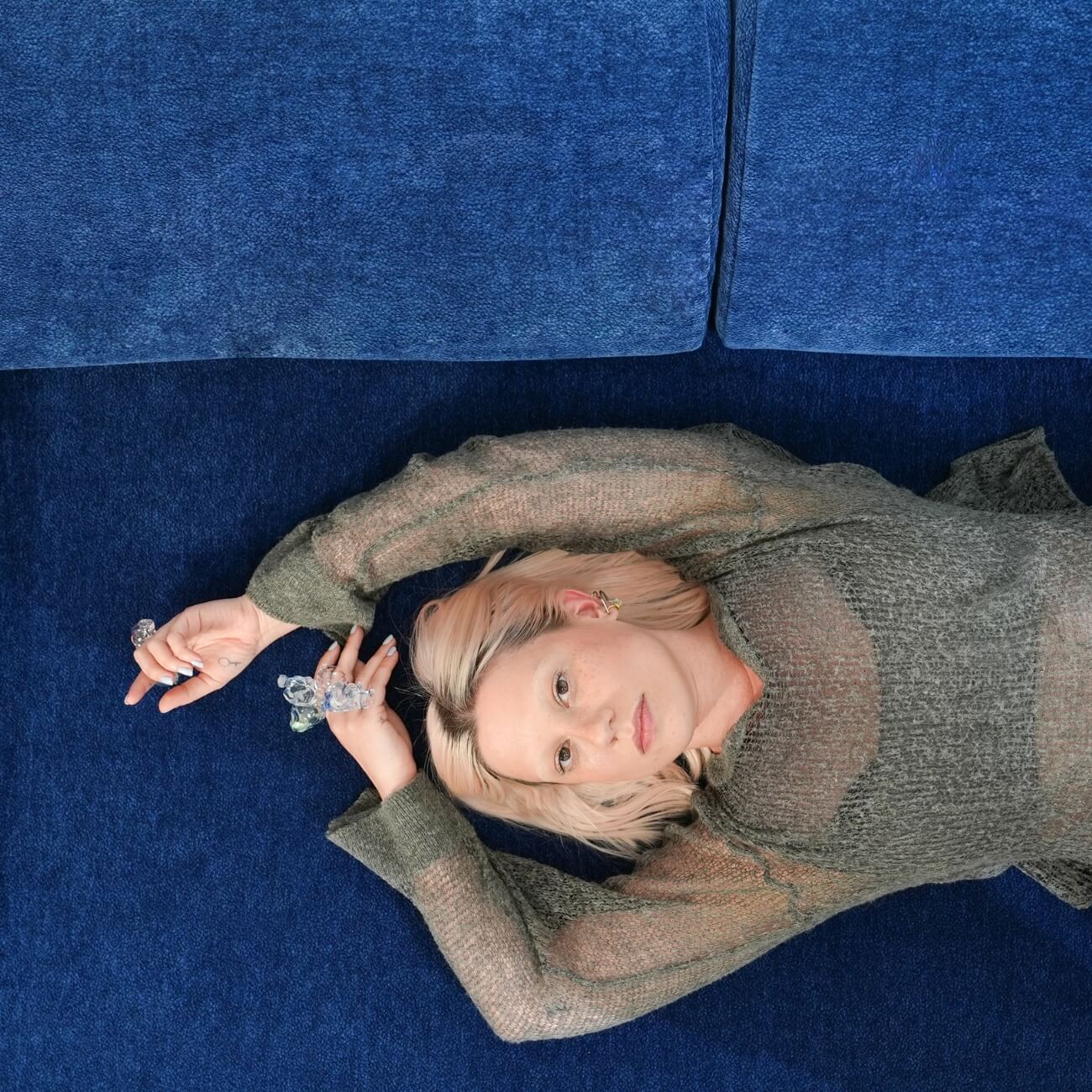 Eine Person mit hellem Haar liegt auf einer blauen Couch und trägt ein halbtransparentes graues Oberteil. Ihre Arme sind über den Kopf gehoben und angewinkelt, und sie trägt Zierringe an den Fingern. Sie blickt ruhig in die Kamera.