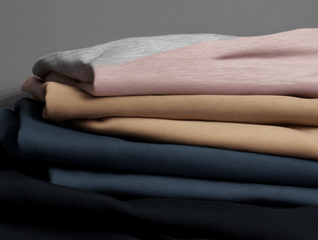 Ein ordentlich gefalteter Stapel Mako-Trikotkleider in verschiedenen Farben, darunter hellgrau, rosa, beige, blau und schwarz, vor einem neutralen Hintergrund.