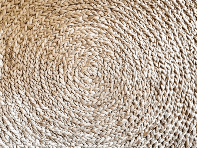 Nahaufnahme einer gewebten Sisalstrohmatte, die ein kreisförmiges, spiralförmiges Muster mit eng verflochtenen, naturfarbenen Fasern in verschiedenen Beigetönen zeigt. Die Textur wirkt rau und handgefertigt.