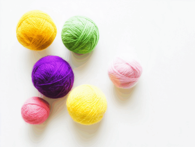 Sechs Wollknäuel in den Farben Gelb, Grün, Lila, Rosa und Hellrosa sind auf einer weißen Fläche angeordnet. Die Garnknäuel sind unterschiedlich groß und liegen dicht beieinander.