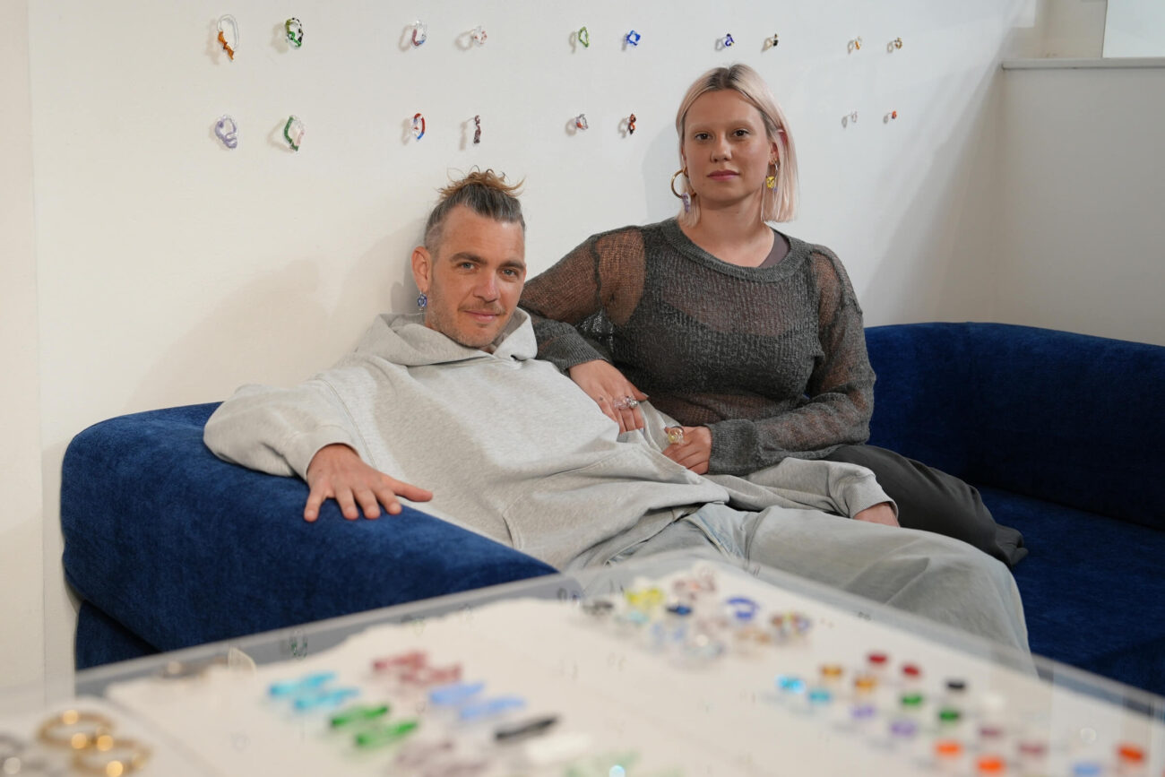 Zwei Personen sitzen auf einer blauen Couch, die eine in einem grauen Kapuzenpulli, die andere in einem dunklen Netzoberteil, zurückgelehnt. Im Vordergrund sind bunte Schmuckstücke ausgestellt und an der weißen Wand hinter ihnen angebracht.