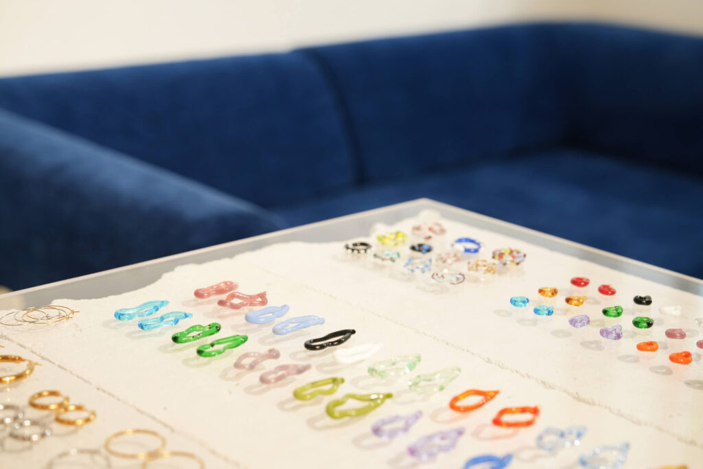 Im Vordergrund steht eine Vitrine mit bunten Ringen, Ohrringen und anderem Schmuck, während im Hintergrund eine blaue Couch verschwimmt.