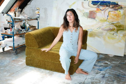 Eine Frau in einem hellblauen Overall sitzt barfuß auf einem grünen Sofa in einem geräumigen Kunststudio, in dessen Hintergrund abstrakte Gemälde und Kunstzubehör zu sehen sind.