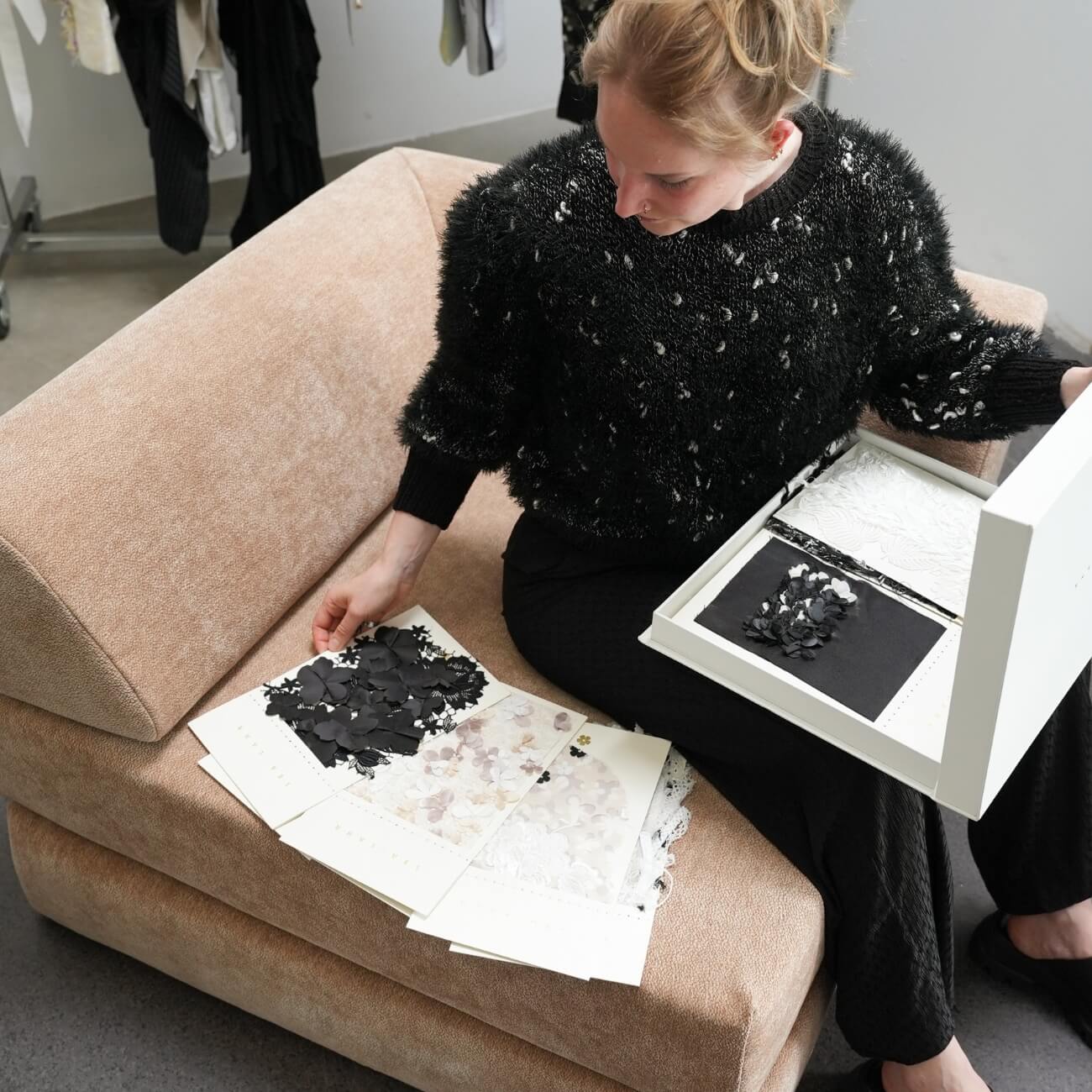 Eine Person, die auf einem beigen Stuhl sitzt, begutachtet Stoffmuster und strukturierte Proben, hält eine Schachtel mit Materialien in der Hand und breitet Musterseiten mit schwarzen und weißen Textilien auf ihrem Schoß aus.