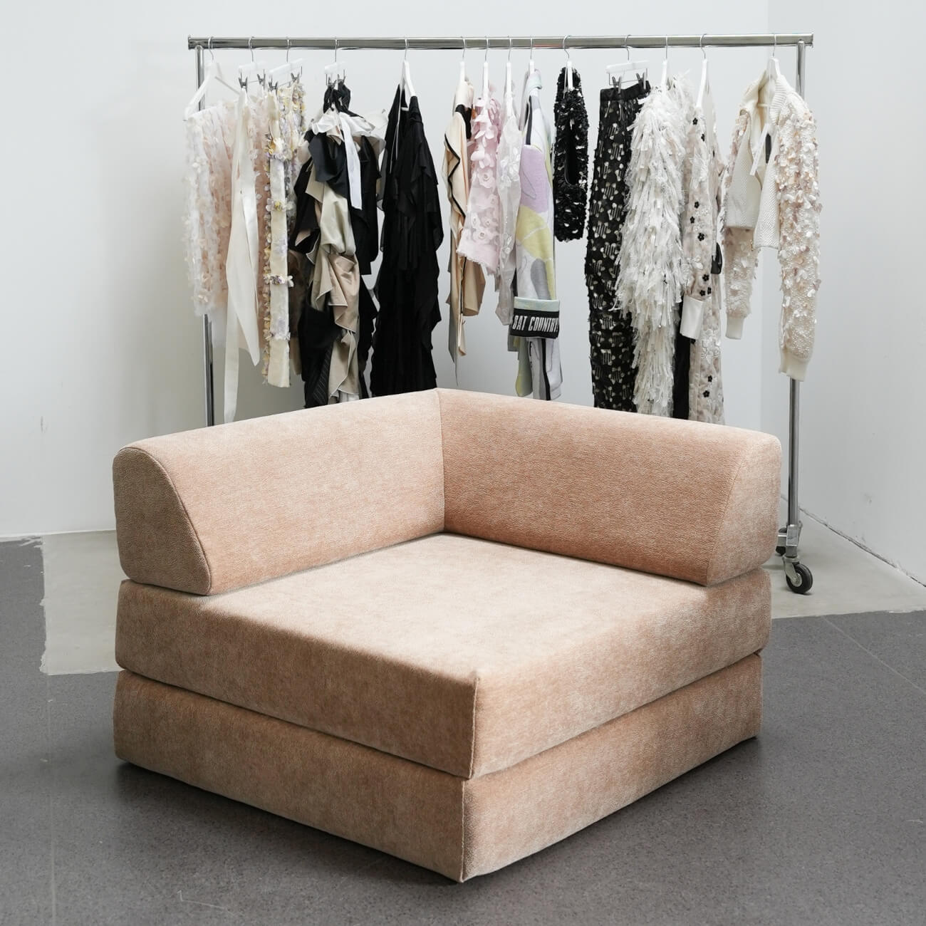 Ein beiger, modern gepolsterter Ecksessel steht auf einem grauen Boden. Dahinter befindet sich ein Kleiderständer mit verschiedenen strukturierten, neutralfarbenen Kleidungsstücken, darunter Rüschen, Fransen und gemusterte Stücke.