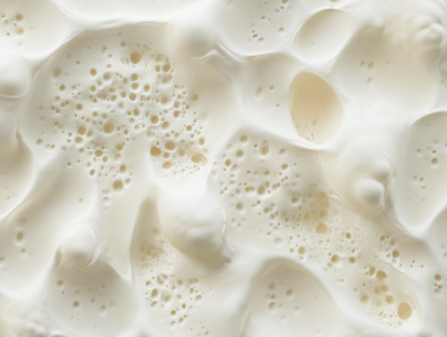 Nahaufnahme einer schaumigen, cremeweißen Latexoberfläche mit verschiedenen Blasen und kleinen Kratern, die der Textur von Schlagsahne oder schaumigem Schaum ähnelt.
