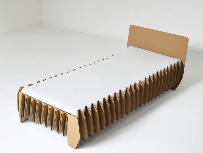 Ein minimalistisches Bett mit weißer Matratze ruht auf einem einzigartigen, von einem Pappbett inspirierten Rahmen aus gefaltetem, ziehharmonikaartigem Karton in einem schlichten, weißen Raum.