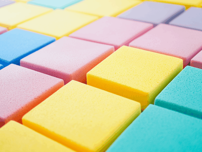 Nahaufnahme von bunten rechteckigen Schaumstoffschwämmen, die in einem ordentlichen Gittermuster angeordnet sind. Die Schwämme in Pastelltönen wie Gelb, Rosa, Blau und Lila sorgen für einen fröhlichen und verspielten visuellen Effekt.