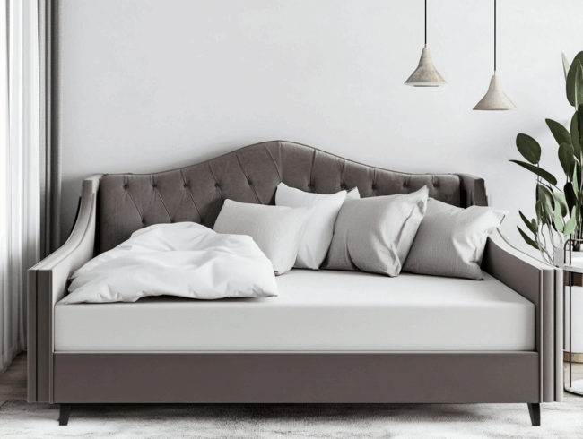 Ein modernes graues Tagesbett mit getufteter Rückenlehne, weißem Bettzeug und mehreren weißen und grauen Kissen steht in einem hellen Raum mit zwei Hängelampen und einer großen grünen Pflanze in der Ecke.
