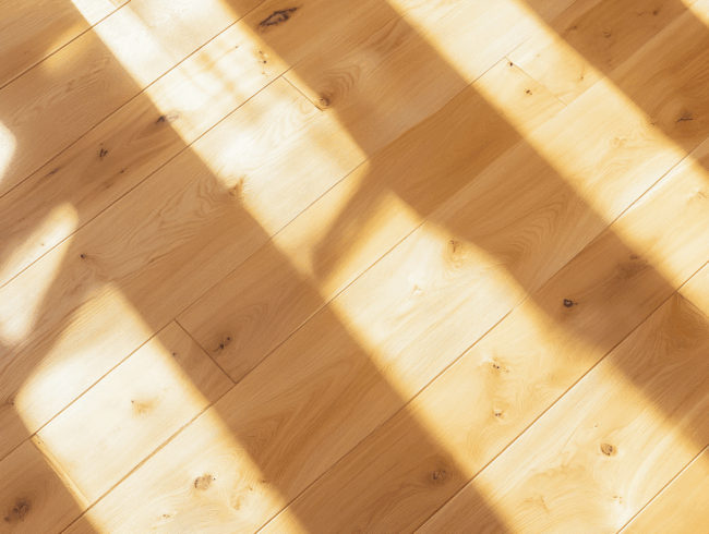 Nahaufnahme eines hellen Holzbodens mit sichtbarer Maserung und Ästen, möglicherweise in Kombination mit Akzenten aus Nussbaumholz, beleuchtet von hellem Sonnenlicht, das von einem Fenster oder einer ähnlichen Struktur einfällt und deutliche rechteckige Schatten wirft.