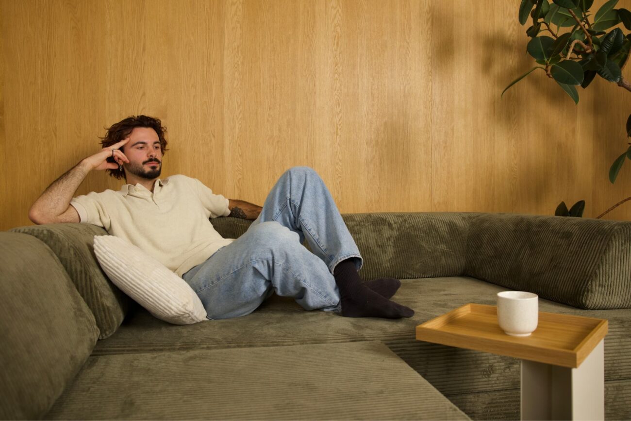 Ein Mann mit lockigem Haar und Bart sitzt lässig auf einem grünen Sofa an einer Holzwand und stützt seinen Kopf auf eine Hand. Ein weißer Becher steht auf einem kleinen Holztisch in der Nähe, eine Pflanze steht in der Ecke.