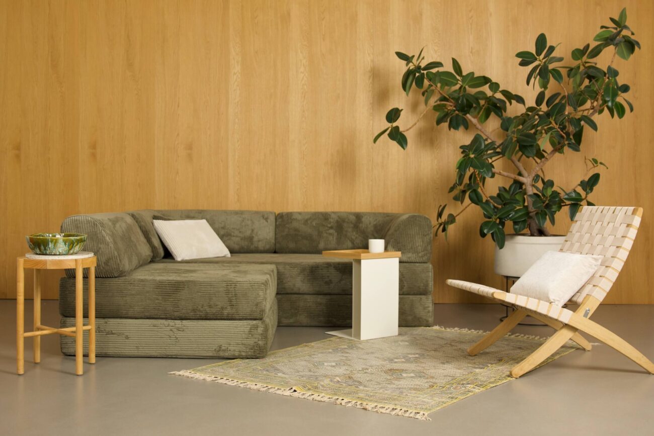 Ein modernes Wohnzimmer mit einem grünen L-förmigen Sofa, einem hellen Holzstuhl, einem runden Beistelltisch mit einer grünen Schale, einem kleinen weißen Couchtisch, einer Topfpflanze und einem beigen Teppich auf einem grauen Boden vor einer holzgetäfelten Wand.