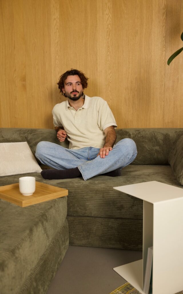 Ein Mann mit lockigem Haar und Bart sitzt im Schneidersitz auf einem grünen Sofa und trägt ein cremefarbenes Poloshirt und eine blaue Jeans. Auf dem Sofa neben ihm steht ein Tablett mit einem weißen Becher, und im Hintergrund ist eine Holzwand zu sehen.