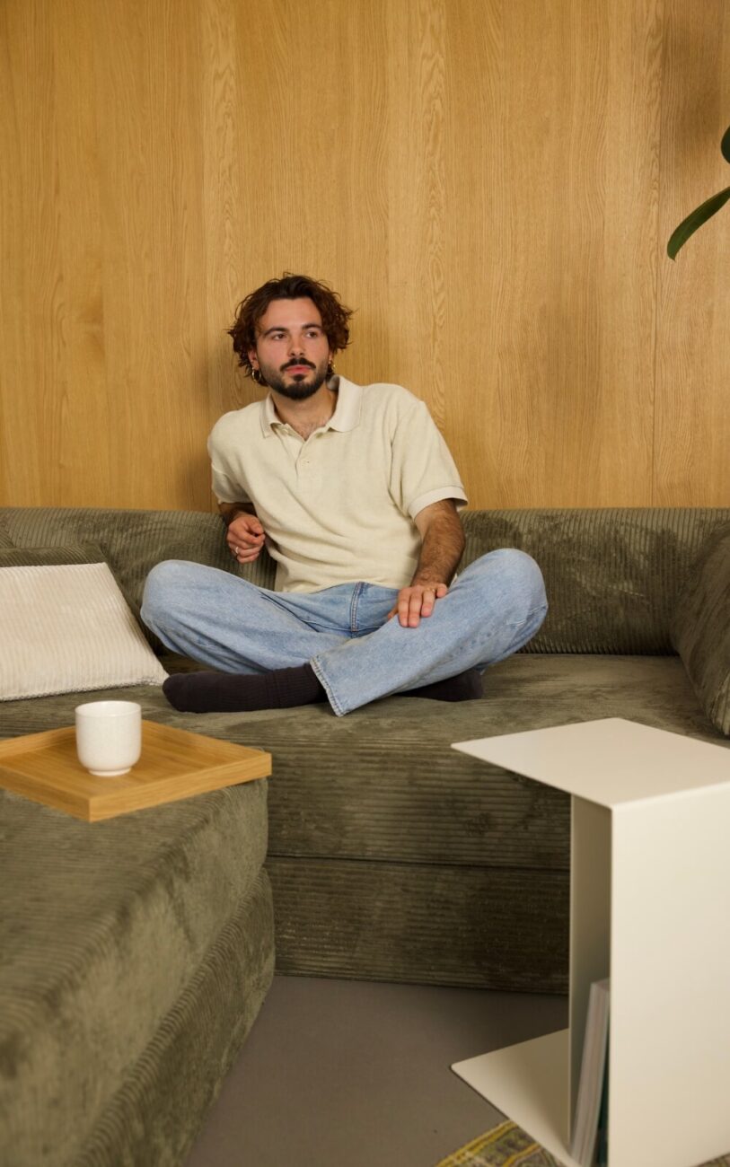 Ein Mann mit lockigem Haar und Bart sitzt im Schneidersitz auf einem grünen Sofa und trägt ein cremefarbenes Poloshirt und eine blaue Jeans. Auf dem Sofa neben ihm steht ein Tablett mit einem weißen Becher, und im Hintergrund ist eine Holzwand zu sehen.