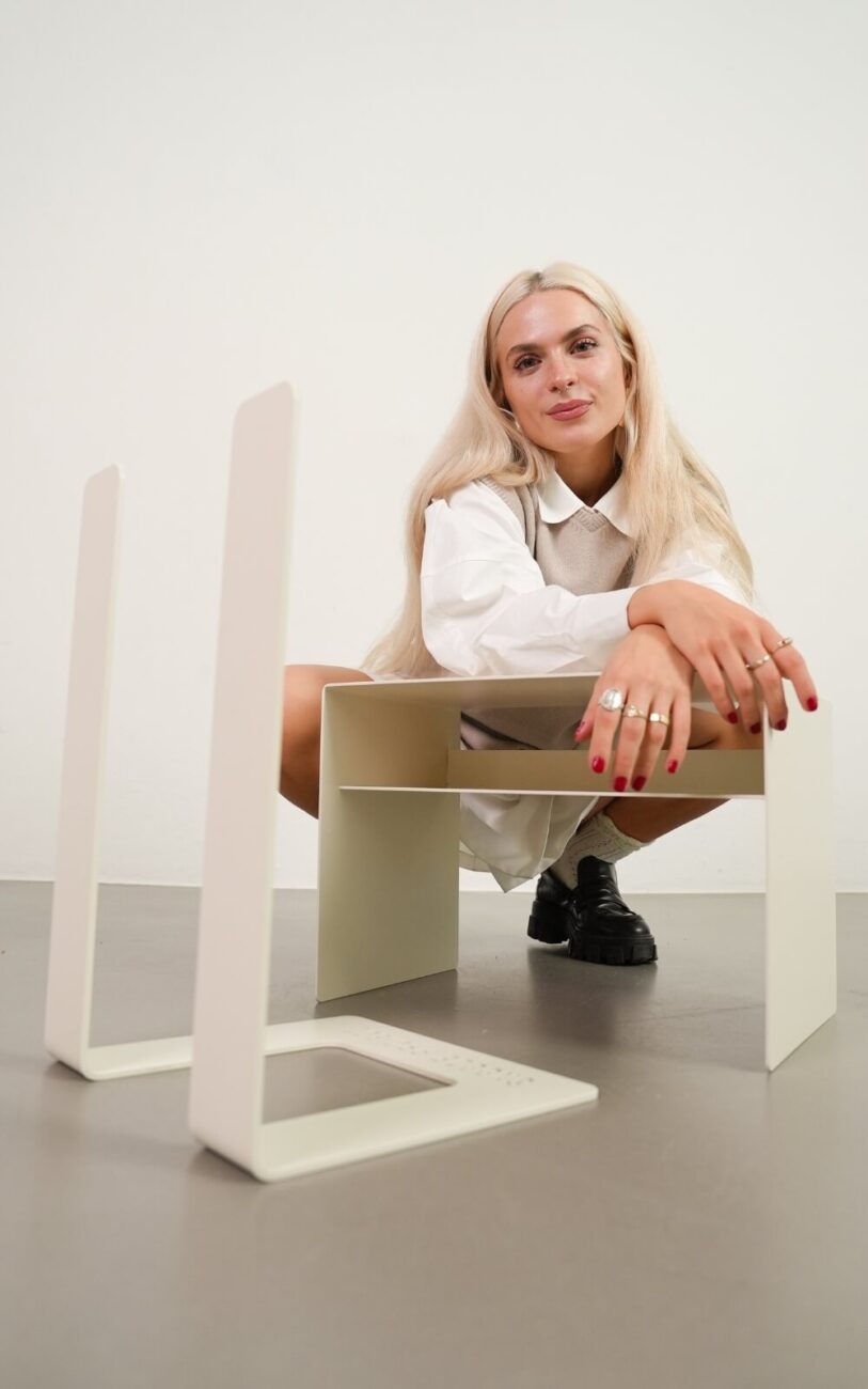 Eine Frau mit langen blonden Haaren hockt hinter einem modernen, minimalistischen Tisch mit hellen Beinen, trägt ein weißes Outfit und schwarze Schuhe in einem hellen Raum mit neutralem Hintergrund.