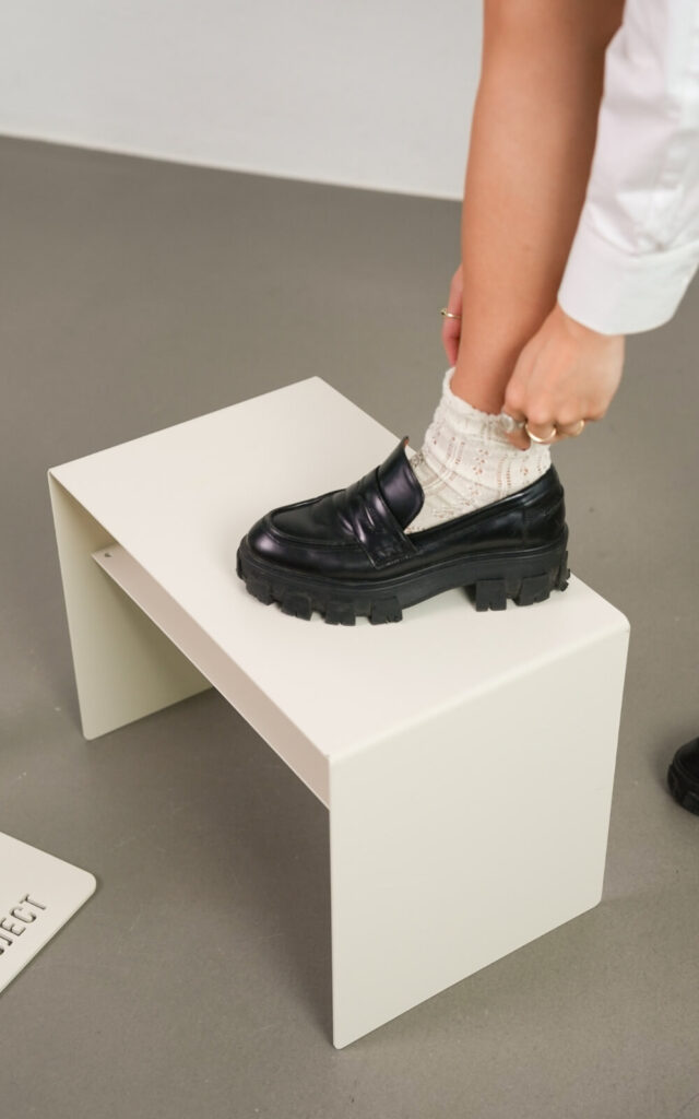 Eine Person steht auf einem kleinen, weißen, rechteckigen Hocker und passt ihre weißen Socken an. Sie trägt einen schwarzen Slipper. Der Boden und der Hintergrund sind grau.